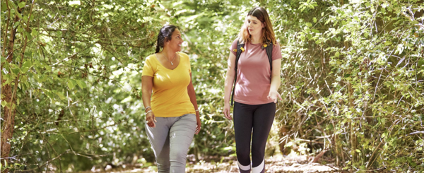 Two women wear exercise clothes and through a forest on a sunny day