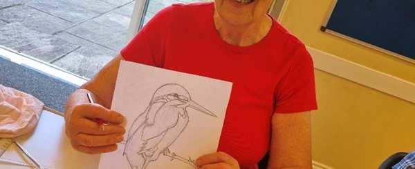 An older woman shows a picture of a kingfisher she has drawn