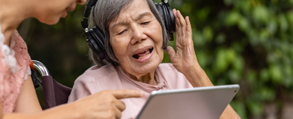 An older Asian women in a wheelchair wears headphones and listens to music on a tablet held up by another woman